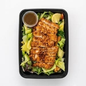 Grilled chicken salad with greens, lemon, and dressing in a black container.